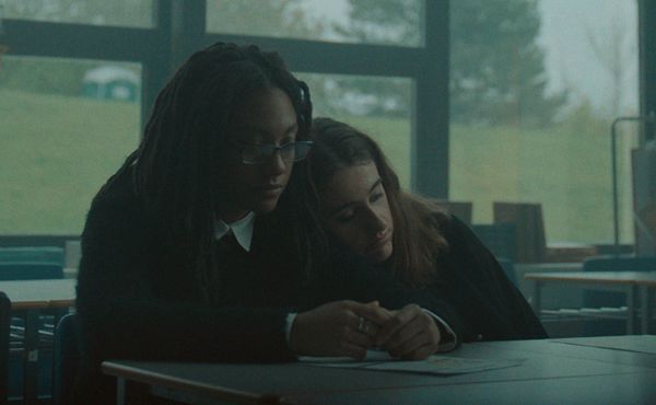 Image from the film 'Tahara' (2020). Two teenage girls sit at connected desks in a classroom, one laying her head on the other’s shoulder. The image has muted colors with dreary, blue-grey lighting washing over the characters and their environment.