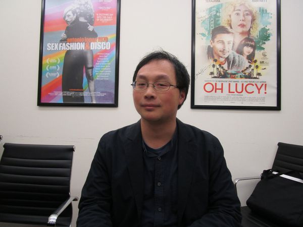 A Girl Missing director Kôji Fukada seated in front of posters for James Crump’s Antonio Lopez 1970: Sex Fashion & Disco and Atsuko Hirayanagi’s Oh Lucy! at Film Movement