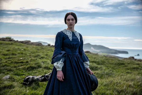 A still from the film Mārama. A young Māori women, dressed in blue Victorian clothes, stands on a grassy clifftop beside the sea.