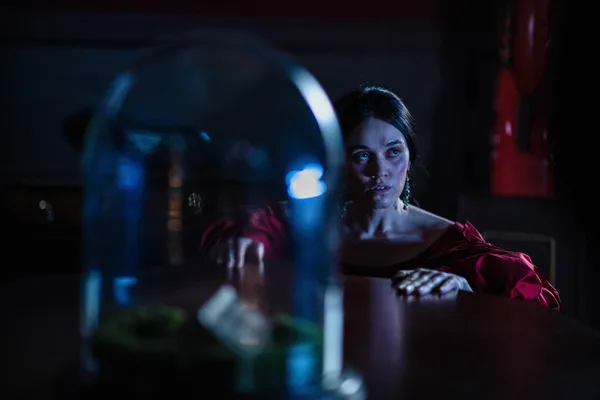 A still from the film Mārama. A Māori woman crouches beside a mahogany table, looking at a bell jar.