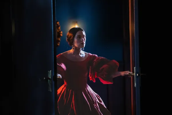 A still from the film Mārama. A young Māori woman in a red dress opens dark blue French doors and peers into a shadowy room.