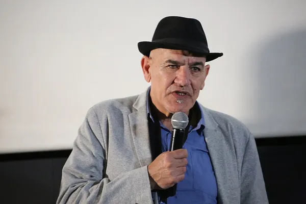 Filmmaker Rashid Masharawi. A middle-aged man wearing a blue shirt, grey jacket and black hat, and holding a microphone.